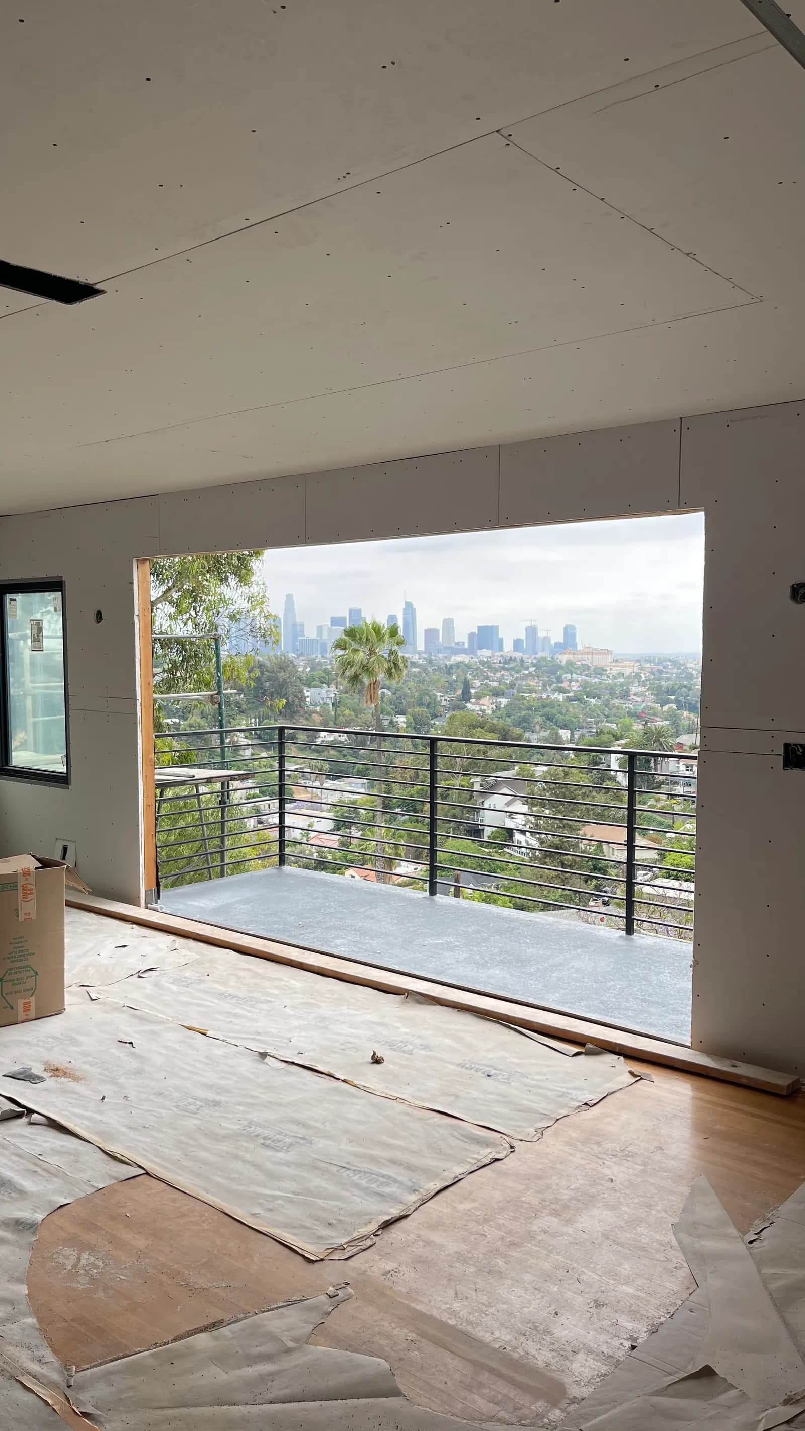 A hillside living room with gypsum board hung and screws set but seams not yet taped, floor protection down, and the downtown Los Angeles skyline through a full-width opening.